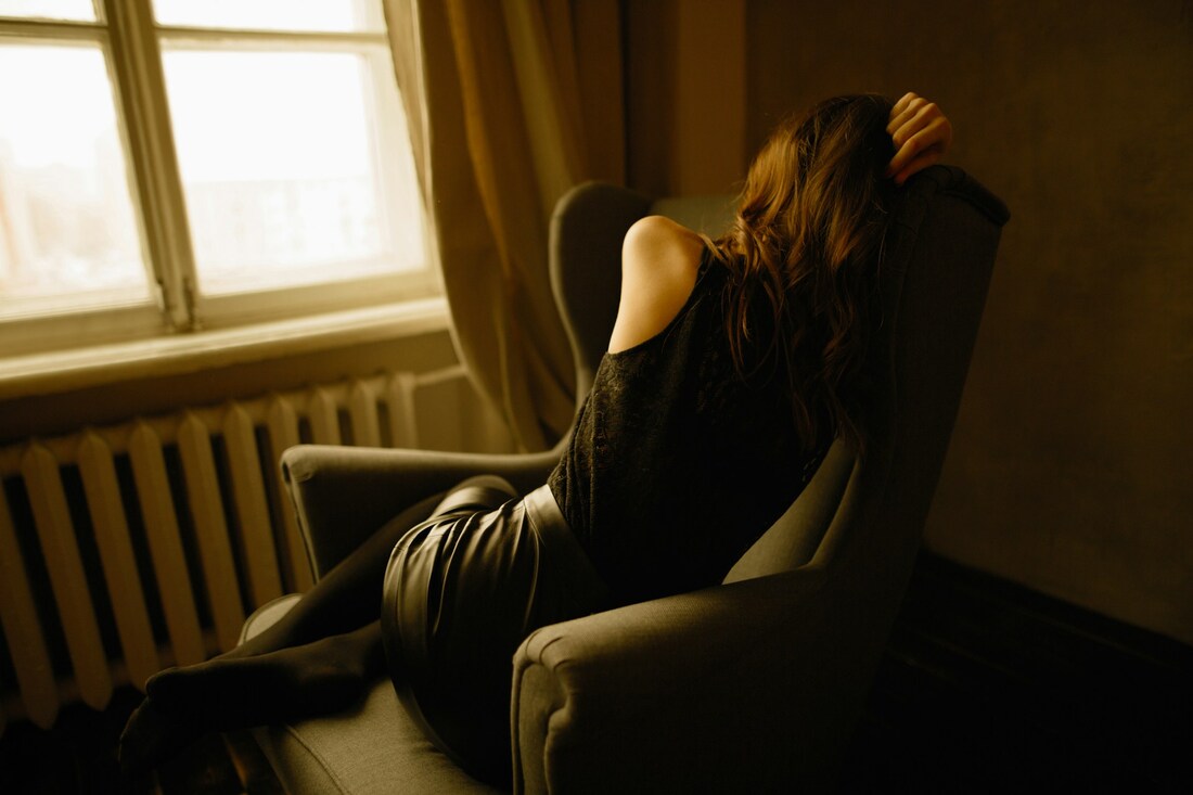 A woman sitting in a chair by a window, looking distressed or overwhelmed.