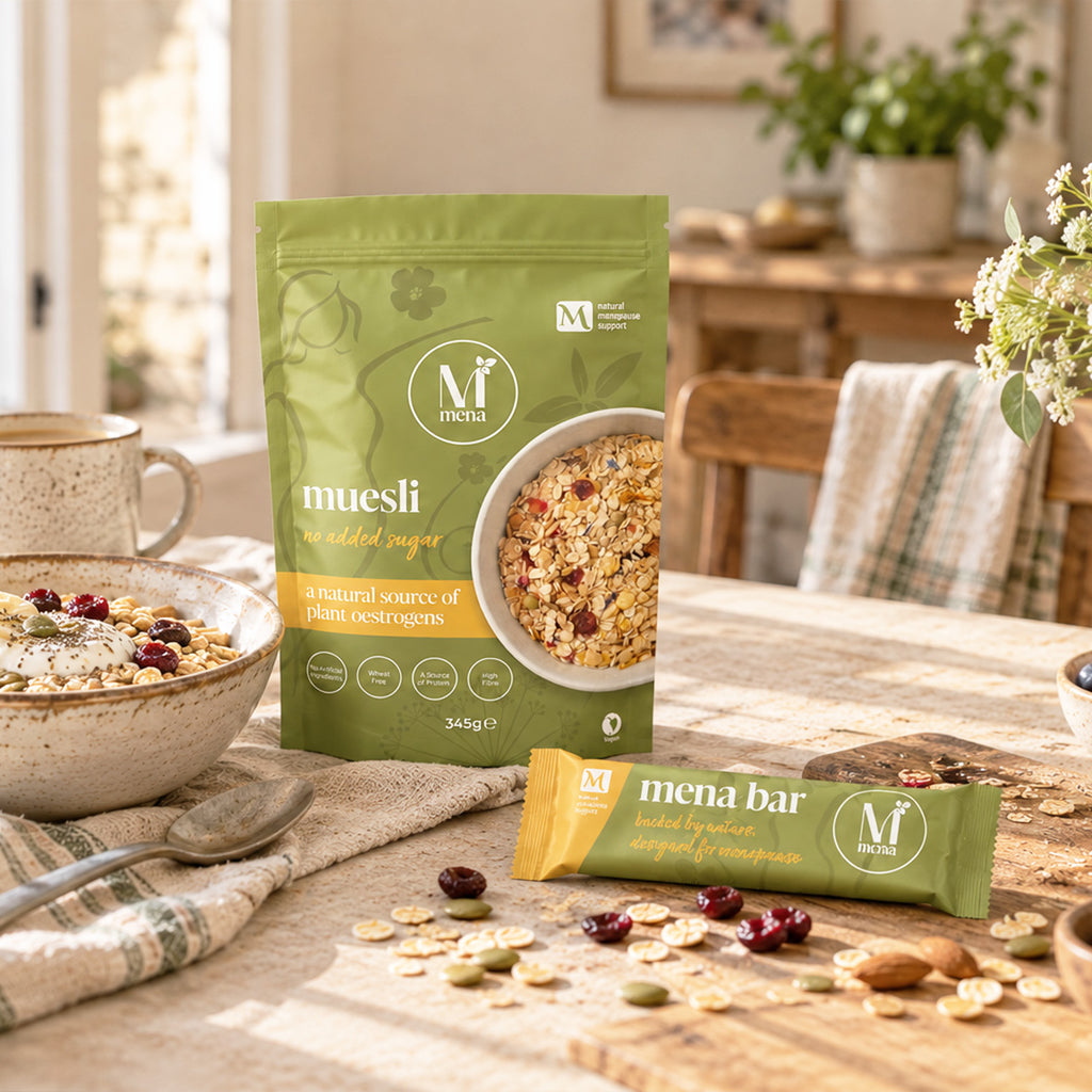 A green stand-up pouch of Mena Muesli and a yellow wrapped Mena Bar are displayed on a rustic wooden table with bowls of oats, dried fruit, and nuts in a warm, sunlit kitchen setting.