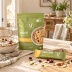 A green stand-up pouch of Mena Muesli and a yellow wrapped Mena Bar are displayed on a rustic wooden table with bowls of oats, dried fruit, and nuts in a warm, sunlit kitchen setting.